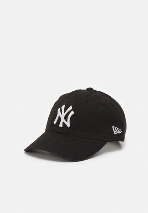 Gorra de béisbol negra con el logo "NY" bordado en blanco en el frente y un logo blanco más pequeño en el lateral, colocada sobre un fondo blanco.