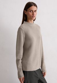 Beiger Strickpullover mit hohem Kragen, langenÄrmeln und strukturiertem Muster. Kombiniert mit dunkel gestreiften Hosen posiert das Modell vor einem neutralen Hintergrund.