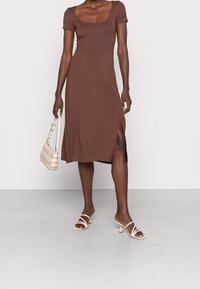 Kvinna i en brun kortärmad midiklänning med sidoklyvning, som håller en liten beige rutig handväska och har vita remsandaler med klack.