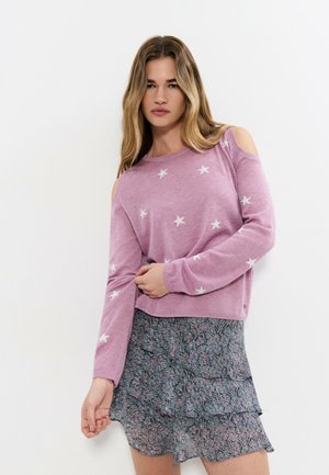 Mujer con un suéter rosa con estrellas blancas y aberturas en los hombros, combinado con una minifalda estampada con volantes, de pie contra un fondo blanco.