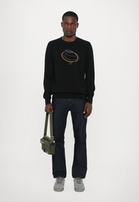 Schwarzes Sweatshirt mit buntem kreisförmigem Grafikdesign, blaue Jeans, graue Sneakers und grüne Umhängetasche mit zwei Reißverschlussfächern.