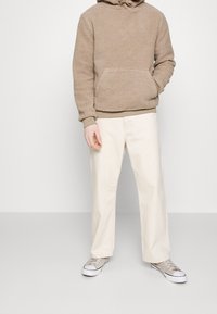 Sudadera con capucha de forro polar marrón con gran bolsillo frontal, combinada con pantalones de pierna ancha color beige claro y zapatillas deportivas grises. Paleta de colores neutros y diseño sencillo.