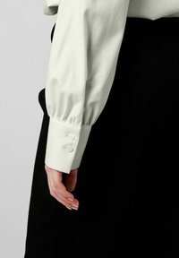 Hellgrüne Bluse mit Puffärmel und Detail mit zwei Knöpfen an den Manschetten, kombiniert mit schwarzem Stoff. Weiche Textur und taillierte Form sichtbar.