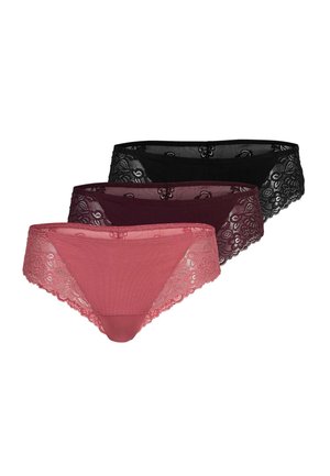 Femme debout pieds nus sur un fond uni, portant un bralette en dentelle rose poussiére assorti et des sous-vêtements, regardant légèrement sur le côté.