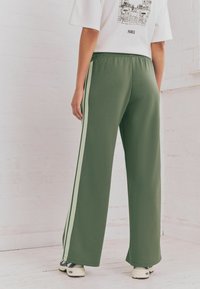 Pantaloni larghi verdi con strisce laterali verde chiaro, vita elastica, materiale in misto cotone, abbinati a una maglietta bianca con grafica e sneakers.