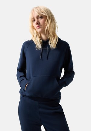Jeune femme aux cheveux blonds portant un sweat à capuche bleu marine et un pantalon assorti, les mains dans la poche de la capuche, expression neutre, fond clair.