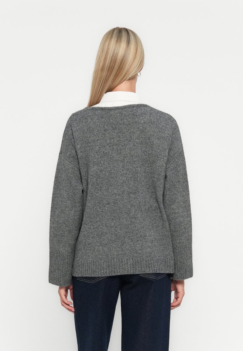 Pull en tricot gris à coupe ample, avec un ourlet côtelé et des manches larges. Superposé à une chemise blanche à col. Texture unie.