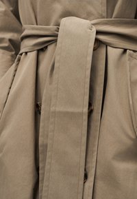 Μπεζ παλτό τύπου trench με υφασμάτινη ζώνη δεμένη, εμφανή κουμπιά και πλαϊνές τσέπες με λεπτομέρειες ραφής.