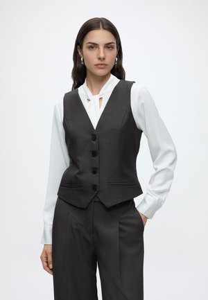 Femme portant un chemisier blanc à manches longues avec un détail découpé, un gilet noir à boutons et un pantalon noir assorti, debout avec une main dans la poche.