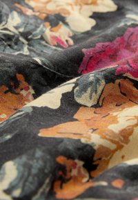 Tissu floral à fond foncé. Présente des fleurs orange, rose et crème avec des feuilles vertes. Surface douce et texturée avec une apparence légèrement froissée.