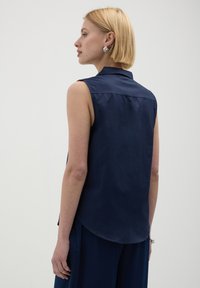 Donna bionda di profilo a sinistra, che indossa una camicia senza maniche di colore blu navy con colletto, pantaloni blu navy coordinati, orecchini d'argento e un braccialetto d'argento.