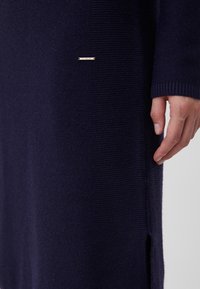 Robe en maille bleu marine foncé, avec une texture côtelée, une fente latérale et une petite plaque dorée près de l'ourlet.