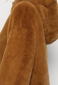 Abrigo de piel sintética marrón con textura suave, que incluye una capucha y un ajuste holgado. La tela tiene un aspecto aterciopelado con un brillo sutil.