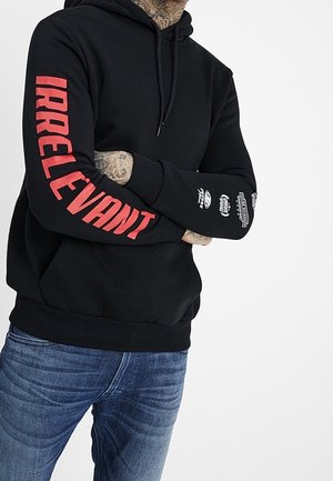 Person med tatoverede hænder iført en sort hættetrøje med "IRRELEVANT" i rødt på det ene ærme og hvide grafikker på det andet, kombineret med jeans.