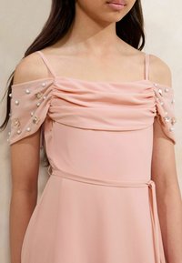 Lichtroze jurk met off-shoulder gedrapeerde mouwen, versierd met bloemapplicaties en embellishments. Soepele, vloeiende stof.