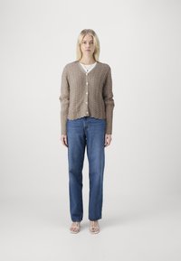 Tan strikket cardigan med kabelmønster, v-hals og bølget kant, kombinert med lyseblå rette jeans og stroppesandaler.