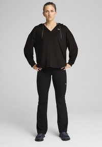 Sudadera negra con capucha y logo blanco, combinada con leggins negros y calzado oscuro. La tela parece suave y un poco holgada.