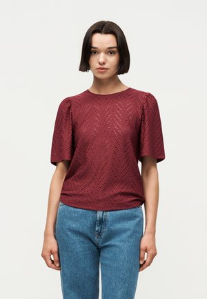 Młoda kobieta z krótkimi ciemnymi włosami, ubrana w bordową teksturowaną bluzkę z krótkim rękawem oraz niebieskie dżinsy, stojąca na tle jednolitego tła.