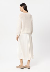 Leichter Pullover aus strukturiertem cremefarbenem Strick, kombiniert mit einem glatten cremefarbenen Rock. Das Modell trägt geflochtene beige Schuhe und ist von hinten gesehen.