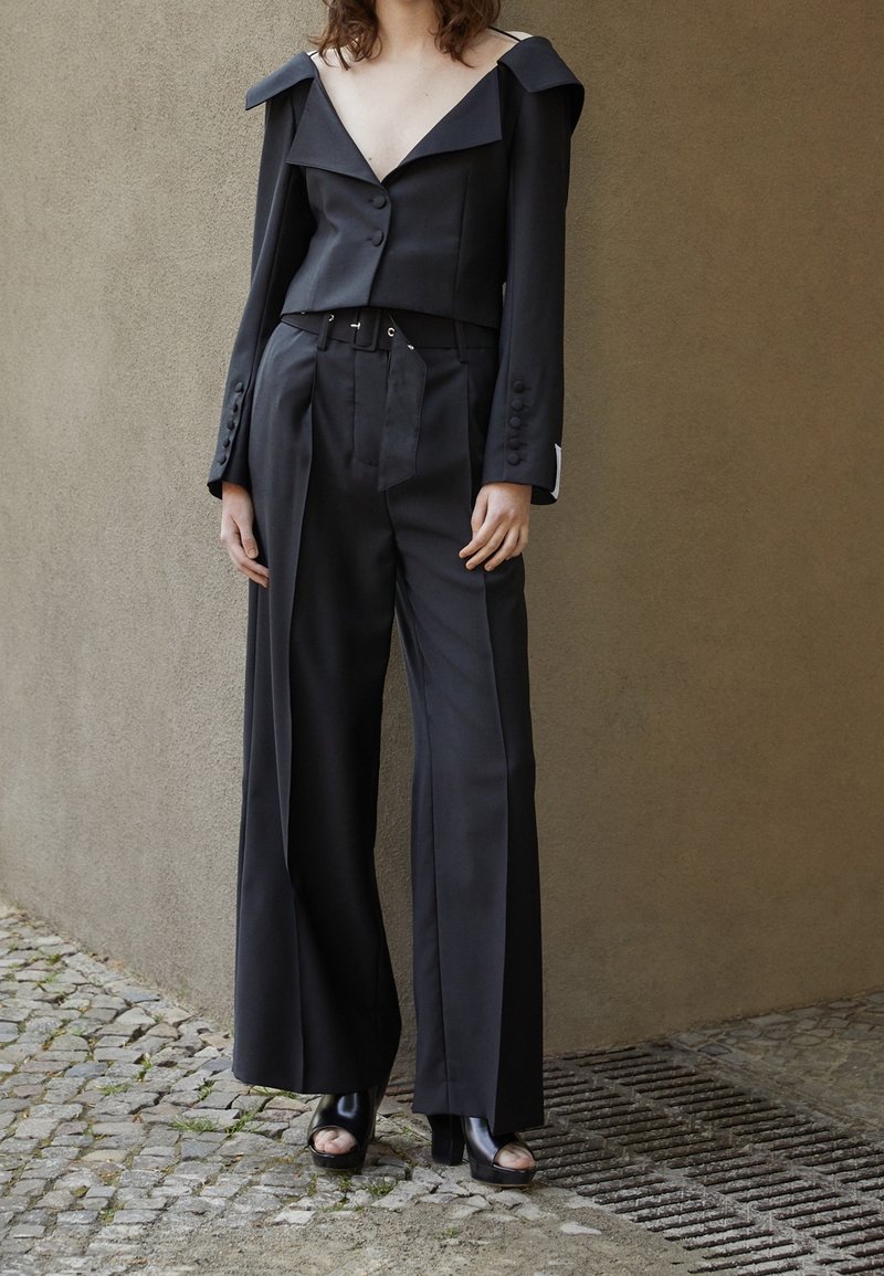 Svart skräddarsydd kostym med en kort, off-shoulder jacka och vida byxor. Har knappar och ett bälte. Slät textur, minimalistisk design.