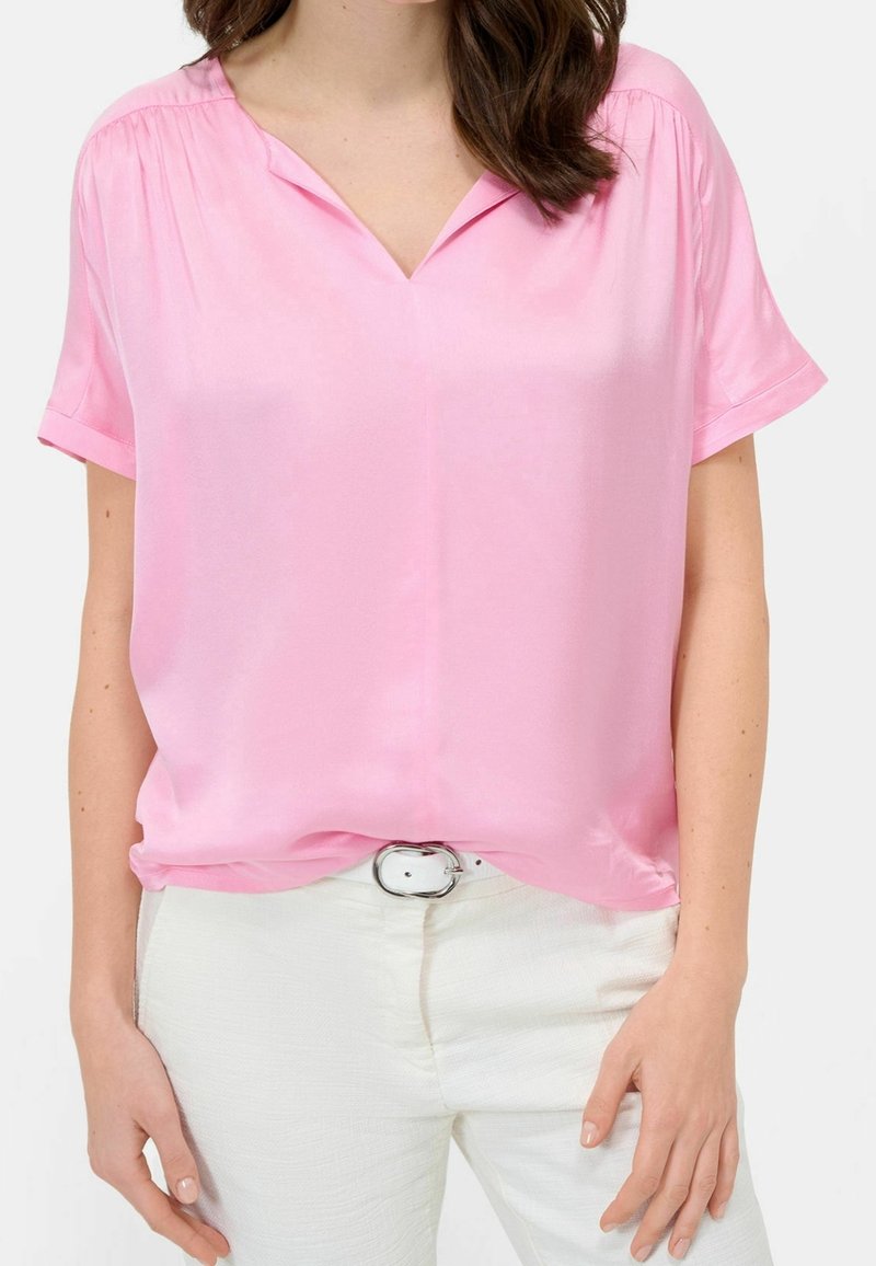 Femme portant un chemisier rose ample à manches courtes, rentrée dans un pantalon blanc avec une ceinture blanche, montrant la partie supérieure du corps du cou jusqu’aux hanches.