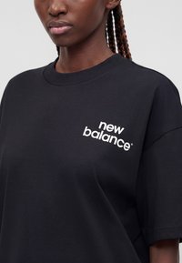 Γυναίκα με πλεξούδες που φοράει μαύρο t-shirt New Balance με λευκό λογότυπο στο αριστερό στήθος, σε απλό φόντο.