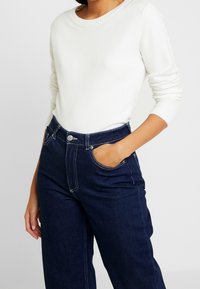 Vit stickad tröja med rund halsringning som matchas med högmidjade mörka jeans i denim med sidofickor och synlig sömnad.