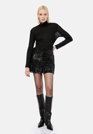 Lyshåret kvinde iført en transparent sort rullekrave, sort paliet-nederdel og knæhøje sorte læderstøvler, stående med hånd på hoften.