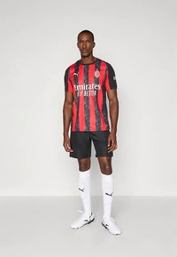 Fotbalový dres s krátkým rukávem červené a černé pruhované barvy, s logem a textem sponzora. Nositý s černými šortkami a bílými ponožkami.