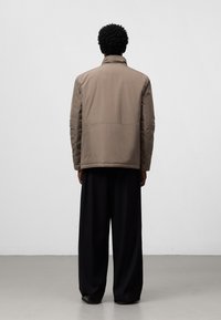 Giacca imbottita marrone con colletto alto, taglio dritto e design minimalista, abbinata a pantaloni neri larghi. Tessuto texturizzato, senza chiusure visibili.