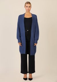 Jaket kardigan panjang rajut berwarna biru tua, dengan tekstur bergaris dan desain depan terbuka. Dipadukan dengan celana jeans hitam dengan pinggang tinggi yang memiliki hiasan halus.