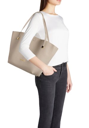 Donna con top bianco e jeans neri porta una grande borsa beige a tracolla, mano in tasca, su sfondo bianco.
