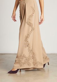 Pantalones de pierna ancha en tela beige con un adornado floral texturizado a lo largo de un lado y dos botones decorativos en la cintura.