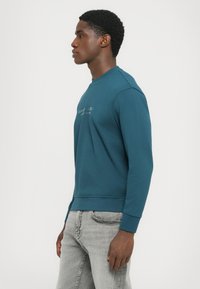 Teal hettegenser med rund hals og ribbede mansjetter. Har en subtil trykt logo på fronten. Modellen har på seg grå jeans.