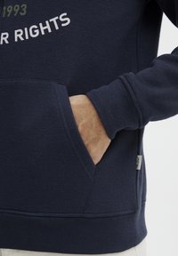 Mornarska majica s kapuco z kenguru žepom, rebrastimi manšeti in kontrastnim belim napisom. Mekano blago z brušenim učinkom.