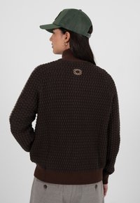 Brun strikket genser med teksturert design, ribbestrikket krage og kant, med en liten logo bak. Brukt med en grønn caps.