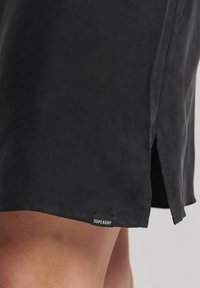 Pantaloni corti neri in tessuto liscio con una fessura laterale, dotati di una piccola etichetta bianca "SUPERDRY" all'orlo.