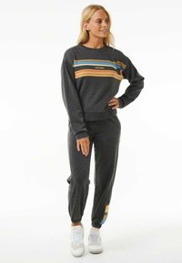 Kvinne iført en mørkegrå hettegenser og matchende joggebukser med flerfarvede striper, som står med en hånd på hoften og hvite sneakers.