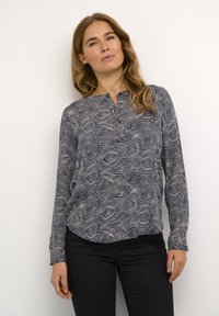 Dlhý rukáv, námornícky modrý blúzka s kontrastným zvlneným bielym vzorom. Obsahuje malý výstrih a voľný strih, vyrobená z mäkkej textílie.