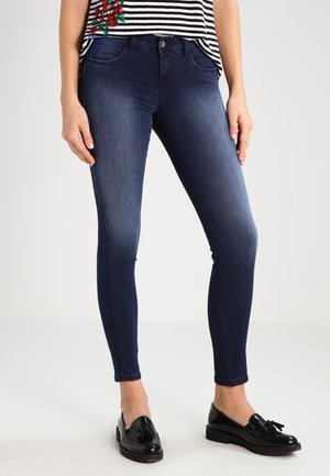 Femme portant un jean skinny bleu foncé, des mocassins noirs à pampilles et un haut rayé noir et blanc avec une broderie florale rouge.
