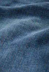 Lähikuva sinisestä teksturoidusta farkkukankaasta, jossa näkyy kudontakuosi ja pehmeitä poimintoja luomassa hienovaraisia varjoja.