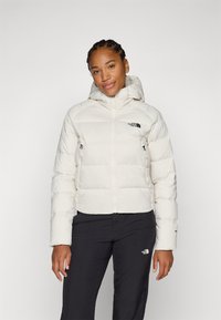 Hvit pufferjakke med hette, med glidelås og sidelommer. The North Face-logoen er vist i svart på brystet.