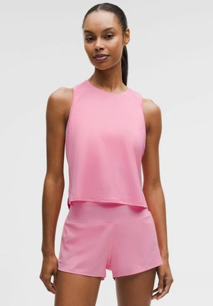 Frau mit dunklem Haar im Pferdeschwanz, trägt ein ärmelloses pinkes Sporttop und passende pinke Shorts, steht vor einem einfarbigen Hintergrund.