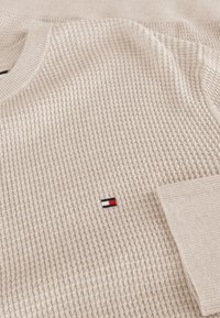 Maglione beige lavorato a maglia con un design testurizzato, caratterizzato da un piccolo patch logo in rosso, bianco e blu sul lato sinistro del petto.