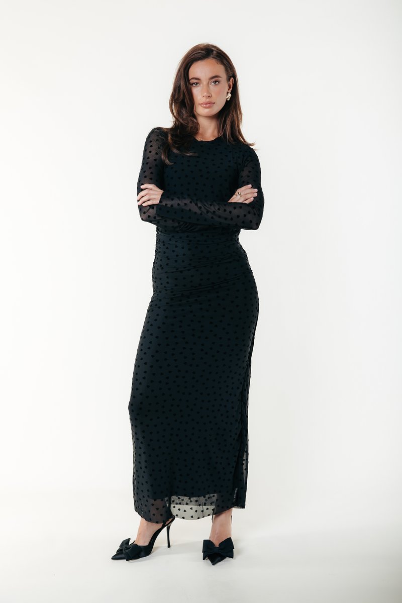 Svart långärmad klänning med figurnära design och prickigt mönster. Den har en sidospjäl och genomskinliga ärmar, kombinerad med svarta klackar med rosett.
