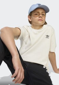 T-shirt crème à manches courtes avec logo Adidas noir, assortie à un pantalon noir avec des rayures blanches. Une casquette bleu clair complète la tenue.