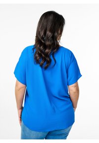 Blå kortærmet bluse lavet af let stof, med en løs pasform og buet kant, parret med lyse denim jeans.