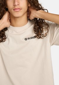 Krémové bavlnené tričko s čiernym logom "ELEMENT" na ľavej strane hrude, s okrúhlym výstrihom a voľným strihom.