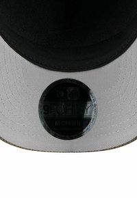 Schwarze Kappe mit einer weißen Unterseite, die einen strukturierten Schirm und einen runden New Era-Aufkleber oben hat, der die Modell- und Anpassungsdetails zeigt.