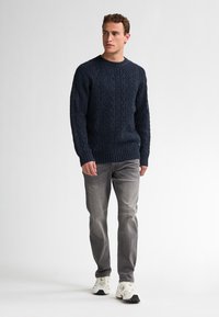 Pulover bleu marin cu tricotaj tip cable și decolteu rotund, cu un model texturat, asociat cu blugi gri deschis și adidași albi. Modelul stă în picioare.